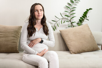 pregnant puzzled woman in gray blouse sits on couch thinking