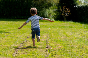 un petit garçon de dos court sur une pelouse en imitant un avion avec ses bras