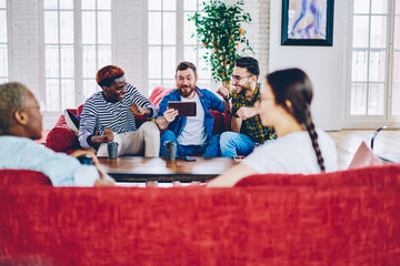 Excited hipster guys dressed in casual wear celebrating victory watching live stream on digital tablet using 4G internet while young women talking during home friendly meeting in modern apartment