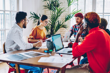 Multicultural group of young people in casual wear discussing plan of collaboration sitting at meeting table, team of diverse creative students cooperating during brainstorming for doing course work