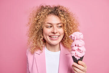 Portrait of cheerful curly haired woman smiles broadly looks at appetizing ice cream in black...