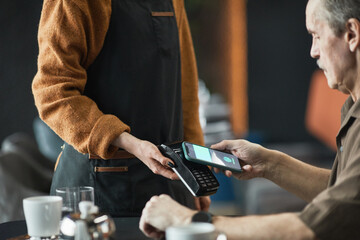 Serious senior man sitting at table and using NFC on smartphone while paying for lunch in cafe,...