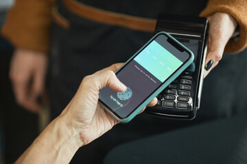 Close-up of unrecognizable customer touching screen to activate app with fingerprint using smartphone while paying by NFC in cafe