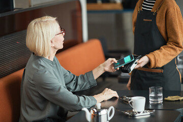 Blond-haired senior Caucasian woman sitting at table in coffee shop and putting smartphone to...