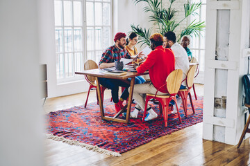 Fototapeta premium Crew of diverse young students learning togetherness during weeknd time in stylish home apartment sitting at table and discussing course work, multiracial hipster guys collaborate on project