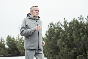 Serious senior Caucasian man in gray sport suit swinging arms while running alone in forest