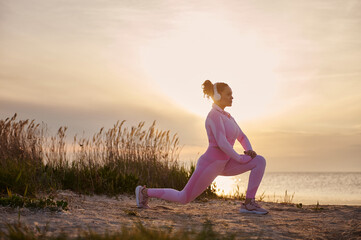 Fototapeta na wymiar Young female athlete doing lunges and warming up at the estuary before an evening run at sunset