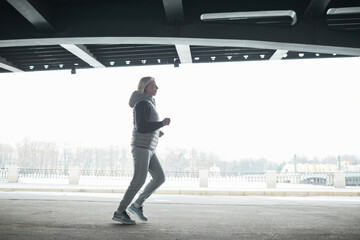 Fit senior woman in gray sport suit jogging alone under bridge while training outdoors