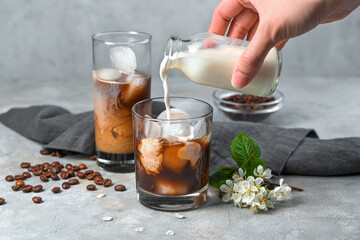 A woman's hand pours milk into a glass of coffee with milk on a gray background.