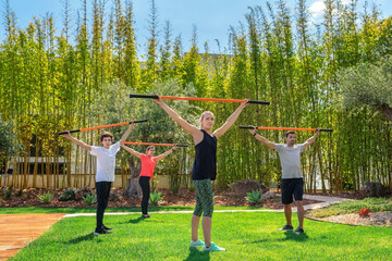 The group is practicing pilate fitness lessons using gymnastic sticks for stretching. In a lovely garden with pool and coach.