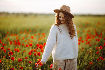 Beautiful woman posing in a poppy field. The girl runs her hand over the poppies. A young woman walks through a flower field with red flowers. Girl in the spring garden. Warm sunset colors.