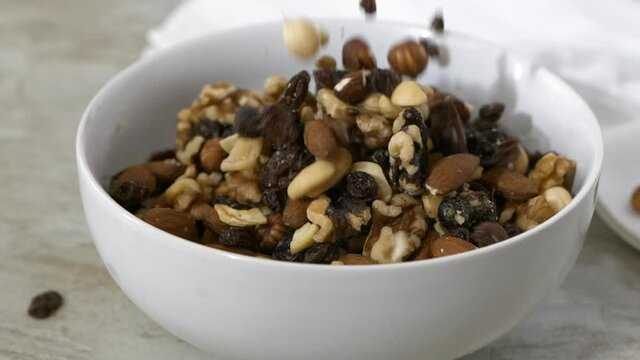Pouring trail mix with almonds, cashews, pecans and raisins into a white bowl slow motion 
