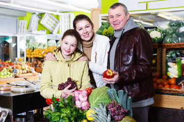 Loving parents and happy daughter with purchases in trolley during family shopping in fruit shop