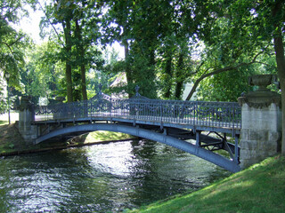 A curved, metal, deserted pedestrian bridge in a park over a river surrounded by green trees and meadows. The sun is reflected between the trees in the flowing water of the river.