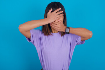 young beautiful asian woman wearing purple t-shirt against blue wall Covering eyes and mouth with hands, surprised and shocked. Hiding emotions.