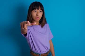 young beautiful asian woman wearing purple t-shirt against blue wall Pointing with finger surprised ahead, open mouth amazed expression, something on the front.