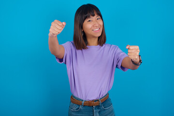 young beautiful asian woman wearing purple t-shirt against blue wall  imagine steering wheel helm rudder passing driving exam good mood fast speed