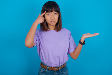 Fototapeta premium young beautiful asian woman wearing purple t-shirt against blue wall confused and annoyed with open palm showing copy space and pointing finger to forehead. Think about it.