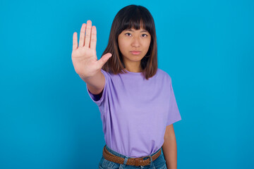 young beautiful asian woman wearing purple t-shirt against blue wall doing stop sing with palm of the hand. Warning expression with negative and serious gesture on the face.