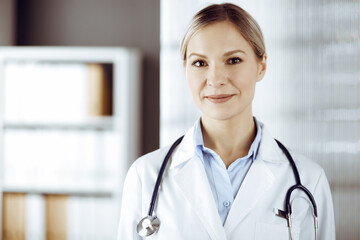 Friendly female doctor standing in sunny clinic. Portrait of cheerful smiling physician. Perfect medical service in hospital