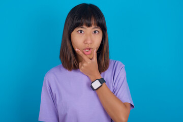 Fototapeta premium young beautiful asian woman wearing purple t-shirt against blue wall Looking fascinated with disbelief, surprise and amazed expression with hands on chin