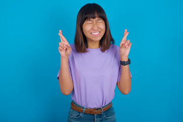 young beautiful asian woman wearing purple t-shirt against blue wall gesturing finger crossed smiling with hope and eyes closed. Luck and superstitious concept.