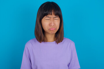Obraz premium Dismal gloomy rejected young beautiful asian woman wearing purple t-shirt against blue wall has problems and difficulties, curves lower lip and closes eyes in despair, being in depression