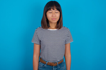 young beautiful asian woman wearing stripped t-shirt against blue wall nice-looking sweet charming cute attractive lovely winsome sweet peaceful closed eyes