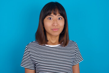 Stunned young beautiful asian woman wearing stripped t-shirt against blue wall stares reacts on...