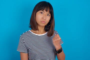 young beautiful asian woman wearing stripped t-shirt against blue wall shows fist has annoyed face expression going to revenge or threaten someone makes serious look. I will show you who is boss