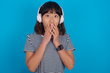 Shocked young beautiful asian woman wearing stripped t-shirt against blue wall stares fearful at camera keeps mouth widely opened wears wireless stereo headphones on ears