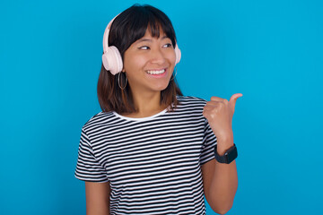 Fototapeta premium young beautiful asian woman wearing stripped t-shirt against blue wall listens audio track via wireless headphones points thumb away advertises copy space
