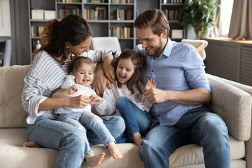 Overjoyed young Caucasian family with two small daughters sit relax on sofa have fun playing game...