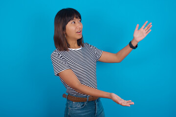 Fototapeta na wymiar Funny astonished young beautiful asian woman wearing stripped t-shirt against blue wall look empty space with arms opened ready to catch something.
