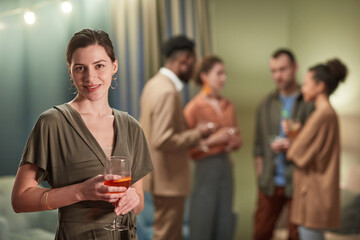 Waist up portrait of elegant young woman looking at camera and holding cocktail glass while enjoying party indoors, copy space