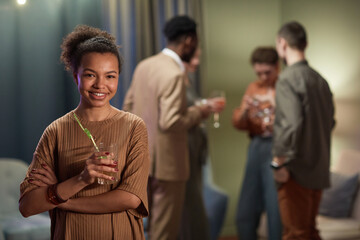 Waist up portrait of smiling mixed race woman looking at camera and holding cocktail glass while enjoying party at home, copy space