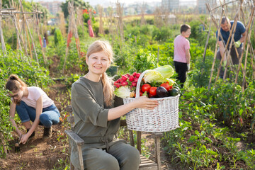 Positive young adult woman posing with basket full of harvested vegetables and greens at homestead, her husband and kids working at garden on background
