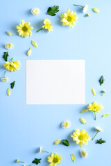 Floral flat lay composition with blank paper card mockup on blue background. Frame of yellow flowers and petals. Top view, overhead.