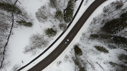 F&uuml;ssen, Germany Bavaria - February 16, 2021: Striped paved winter road winding through the snowy forest trees with a bike path and silver car. Image taken from a drone at 100m altitude. Background.