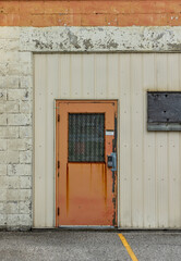 Orange security door against white and orange brick wall and cream-coloured siding. Keyless numbered door padlock and bars on window. Boarded up window. Ontario, Canada.