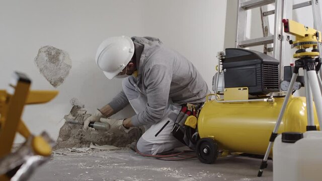 house renovation concept, construction worker wearing helmet at work breaks the old plaster of the wall with pneumatic air hammer chisel, with rubble and tools on background