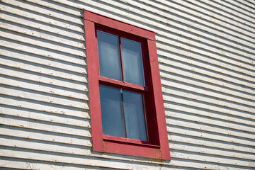 A bright red double hung vintage window with four glass panes. The residential window is on the exterior of a narrow horizontal white wooden clapboard siding wall. The clouds are reflecting in window.