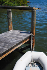 The bow of a small rubber dingy boat or rowboats near a wharf. The boat is white in color. The pier is a small wooden pier surrounded by a pond of blue water. The boat is tied to a wooden post.