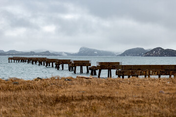 A long dilapidated wharf reaching out to the sea. There are mountains in the background under grey clouds. The ocean is blue with small ripples. There are multiple mountains and fog in the background.