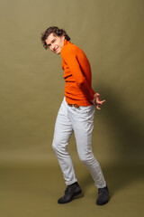Tall handsome man dressed in orange turtleneck and white jeans posing on the green background