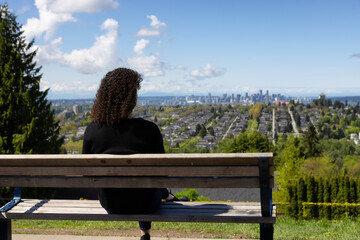 Adult Middle Age Woman sitting outside in a park on the bench and looking at the Modern Cityscape. Burnaby, Vancouver, British Columbia, Canada.