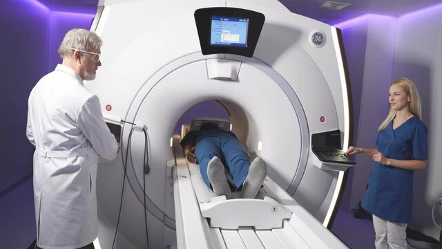 Senior Radiologist Controls MRI or CT or PET Scan with Male Patient Undergoing Procedure. High-Tech Modern Medical Equipment. Friendly Doctor Working With Patient