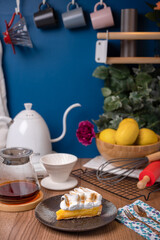 Lemon meringue pie in a black plate on a wooden counter with black coffee, teapot, server, lemon, and baking utensils as a decoration.