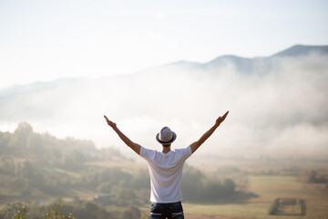 Happy traveler man raising hands up on the top of the world above mountains in white clouds. Hipster guy enjoy amazing atmospheric moment. travel and wanderlust concept