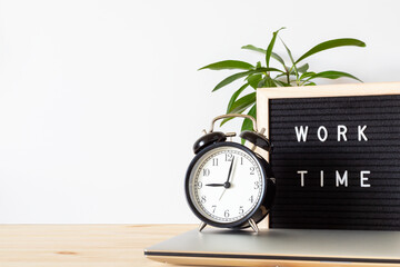 Classic black alarm clock, laptop and black board with words 'work time' on wooden table with white...